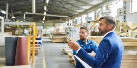 Der Mann in Anzug und weißem Hemd zeigt mit einem Stift selbstbewusst nach links. Ein Arbeiter in blauer Arbeitskleidung blickt entschlossen in die gleiche Richtung. Beide stehen in einer Fabrikhalle. © Adobe Stock, Seventyfour