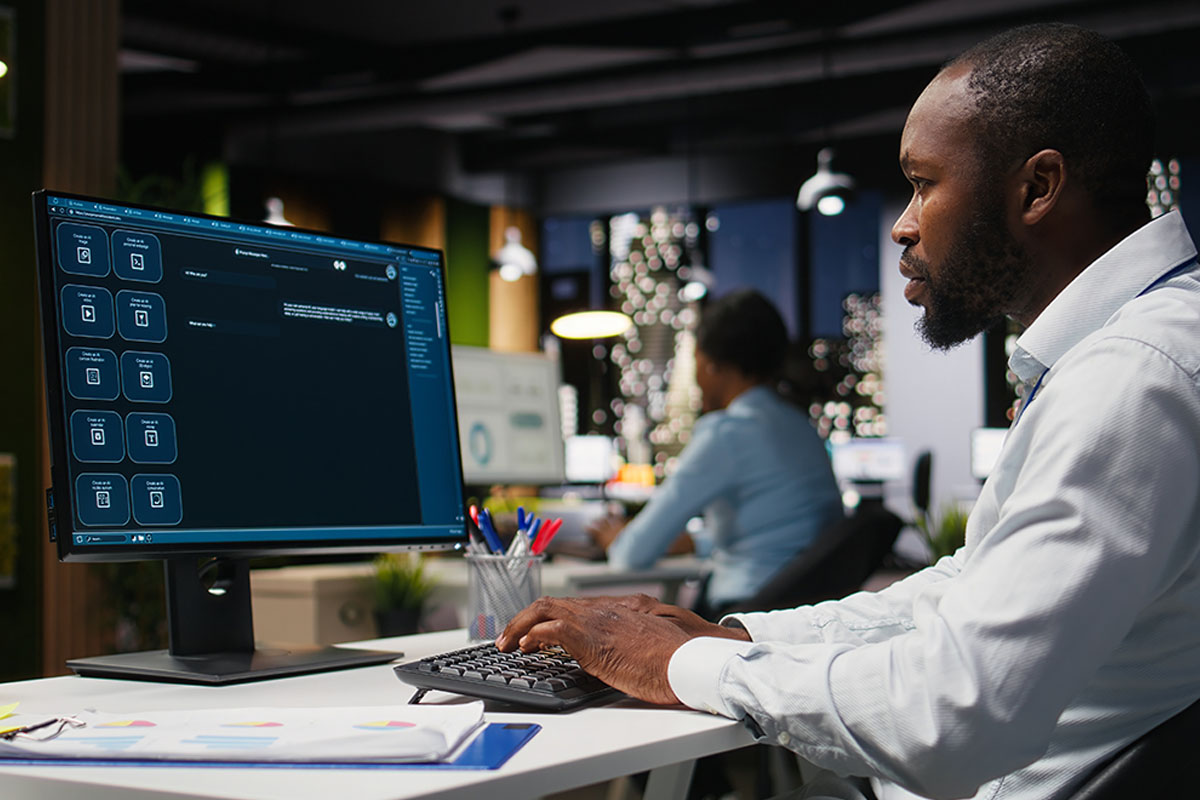 Mann arbeitet am Schreibtisch vor großem Monitor mit Chatbot-Oberfläche im modernen Büro.