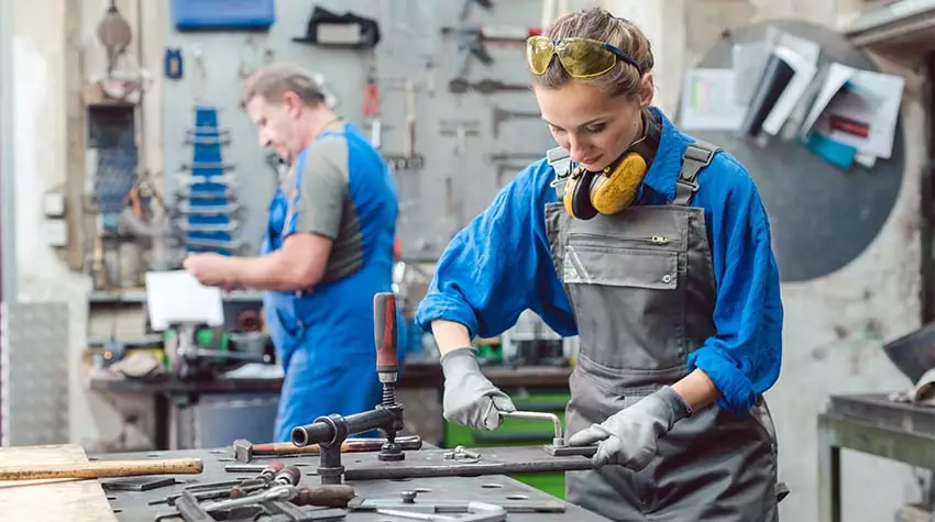 Eine Frau in Arbeitskleidung arbeitet in einer Werkstatt. Hinter ihr arbeitet ein Mann in ähnlicher Kleidung. © Adobe Stock, Kzenon