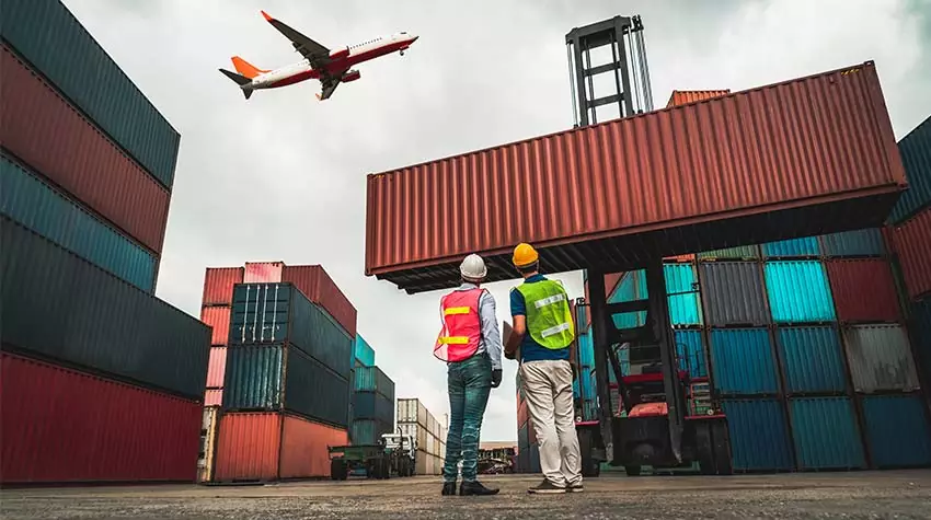 Logistikmitarbeiter stehen zwischen Containern und beobachten ein Frachtflugzeug. © Adobe Stock, Summit Art Creations